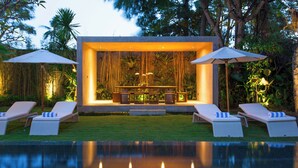 Uma área de estar moderna ao ar livre com piscina, com um espaço central de assentos com mesa e cadeiras, rodeado por espreguiçadeiras e guarda-sóis.