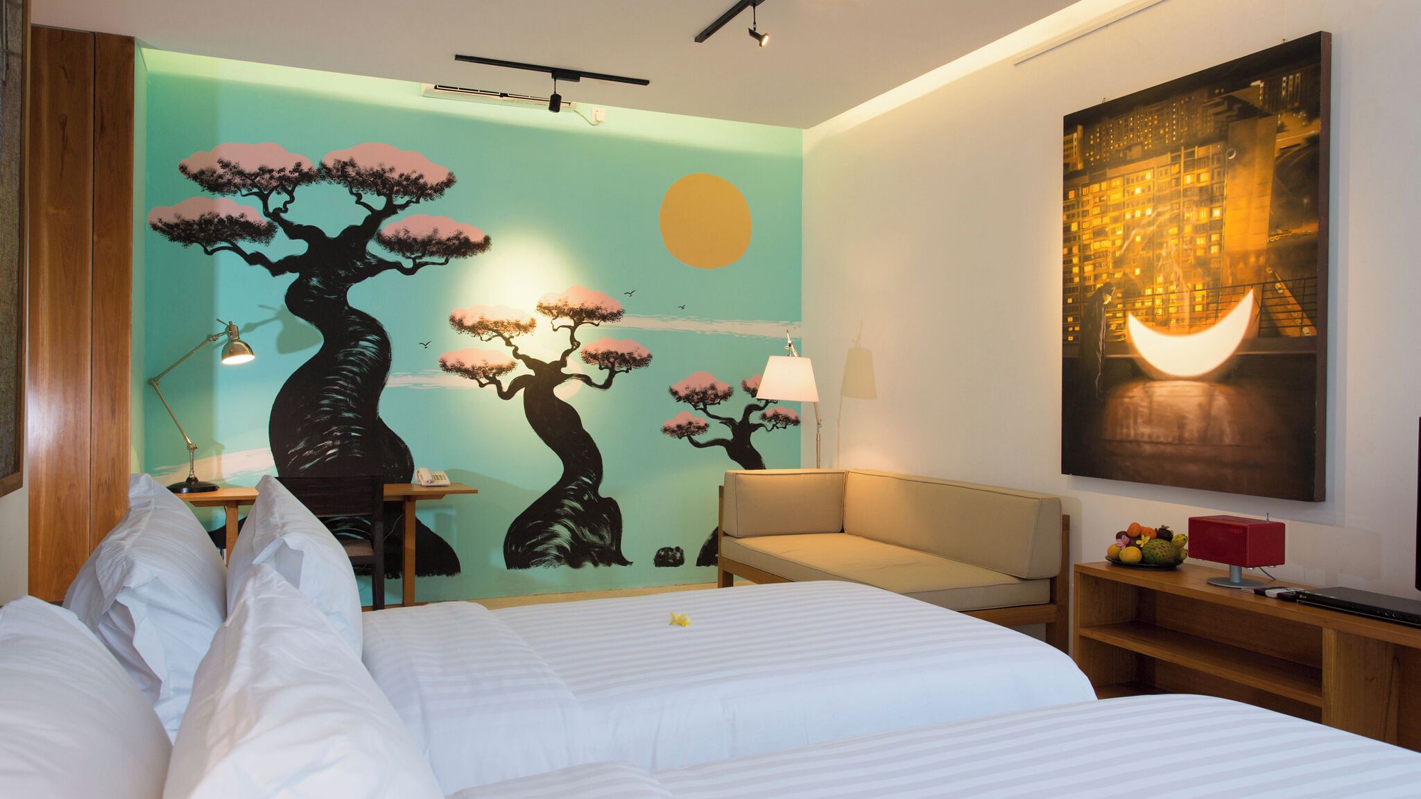 A hotel room with a large bed, a desk with a lamp, a chair, and a wall with a painting of trees and a moon.