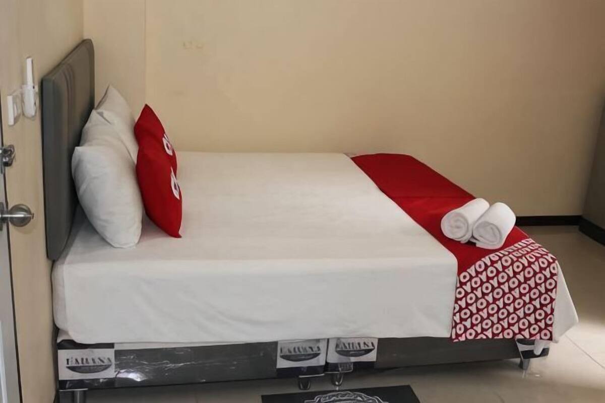 A single bed with a white mattress, red and white pillows, a red blanket, and a patterned throw.