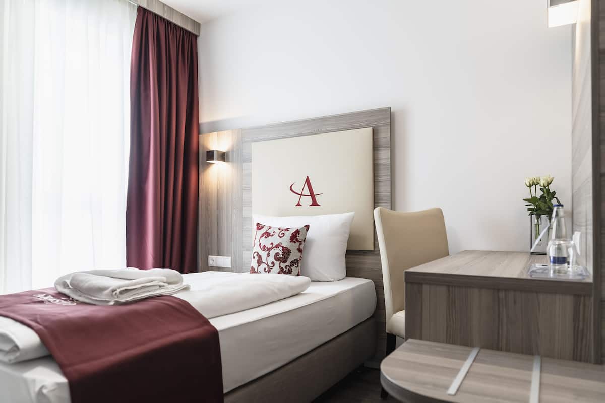 Ett hotellrum med en säng, ett skrivbord, en stol, en vägglampa och en inramad bokstav ’A’ på väggen.