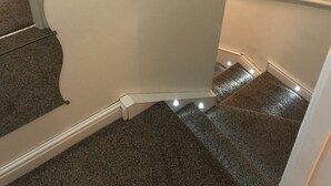 A carpeted staircase with a handrail, leading to a landing with a white wall and a black object on it.
