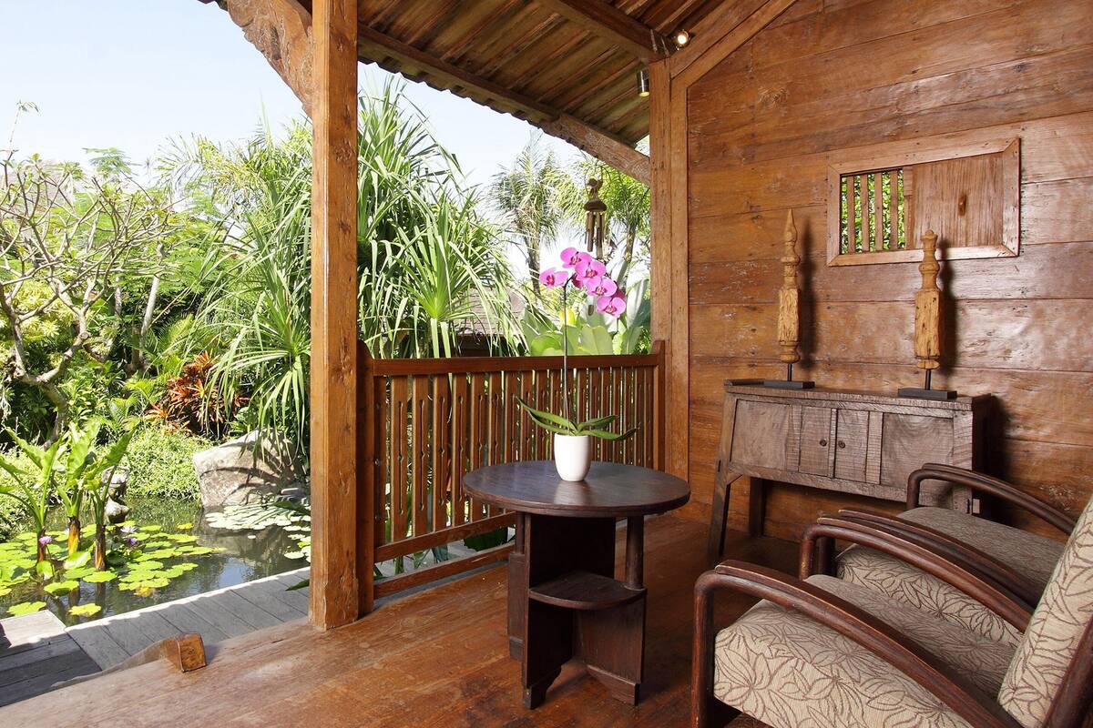 A wooden deck with a table and chairs, surrounded by lush greenery and a small pond.