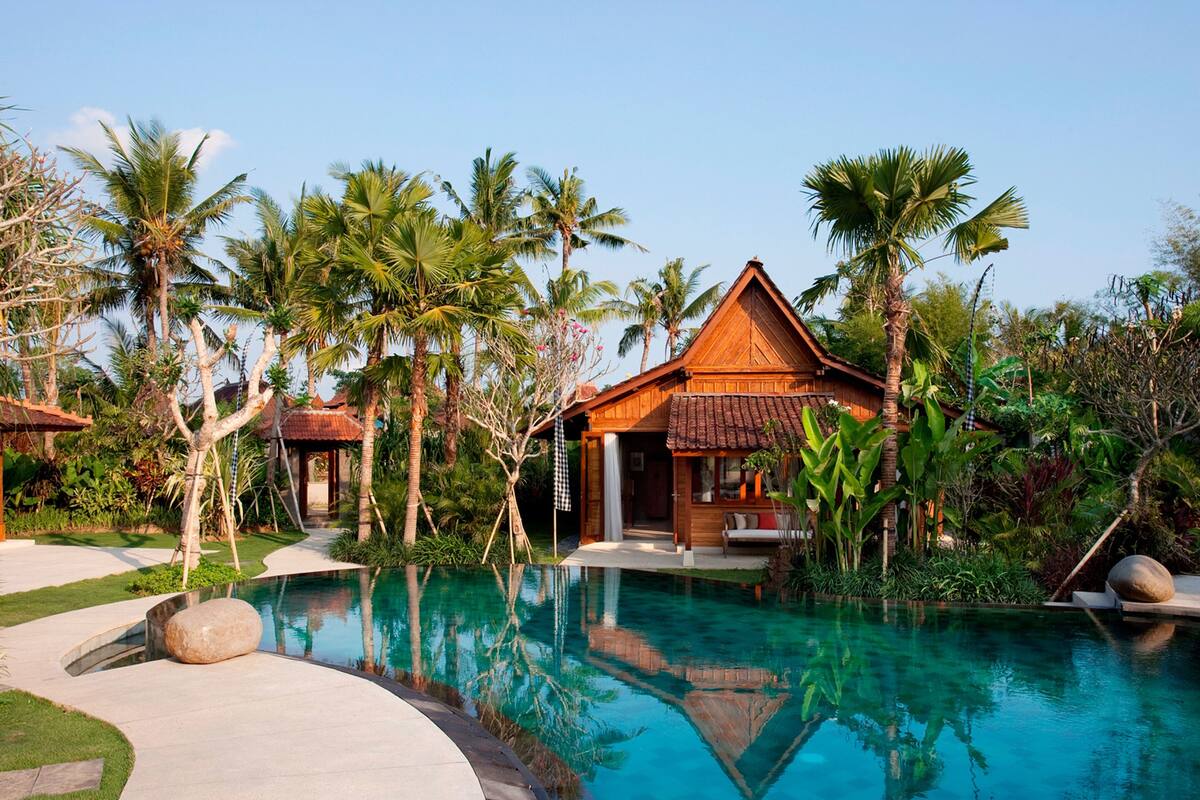 A resort with a swimming pool surrounded by palm trees and a wooden building with a tiled roof.