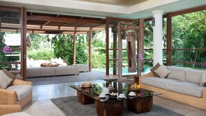 A spacious indoor area with a large window overlooking lush greenery, a wooden ceiling, and comfortable seating arrangements. - Dea Villas (Canggu)