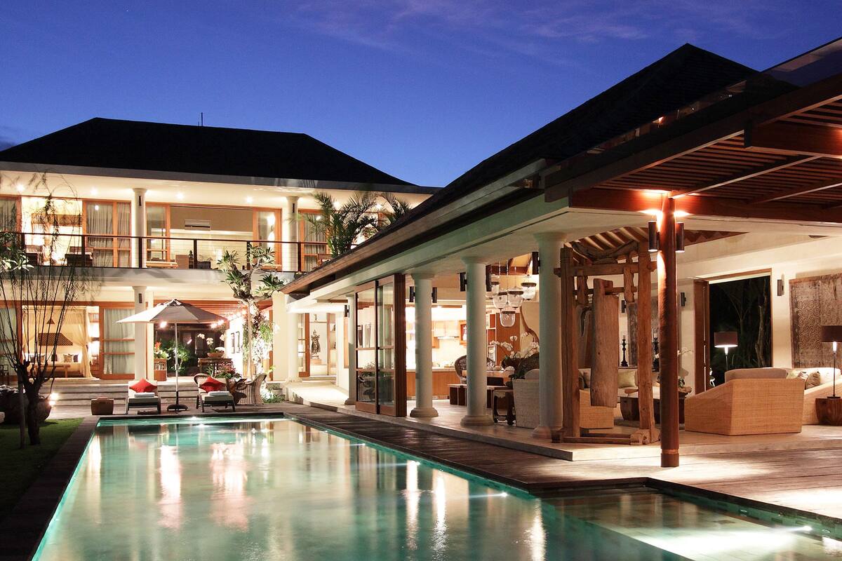 A large, well-lit swimming pool with a covered patio area, surrounded by a two-story building with multiple rooms and balconies.