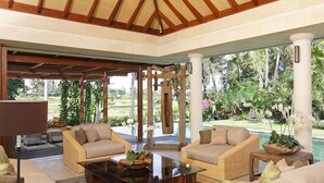 A spacious indoor area with wicker furniture, a glass coffee table, and a view of a lush garden and pool. - Dea Villas (Canggu)