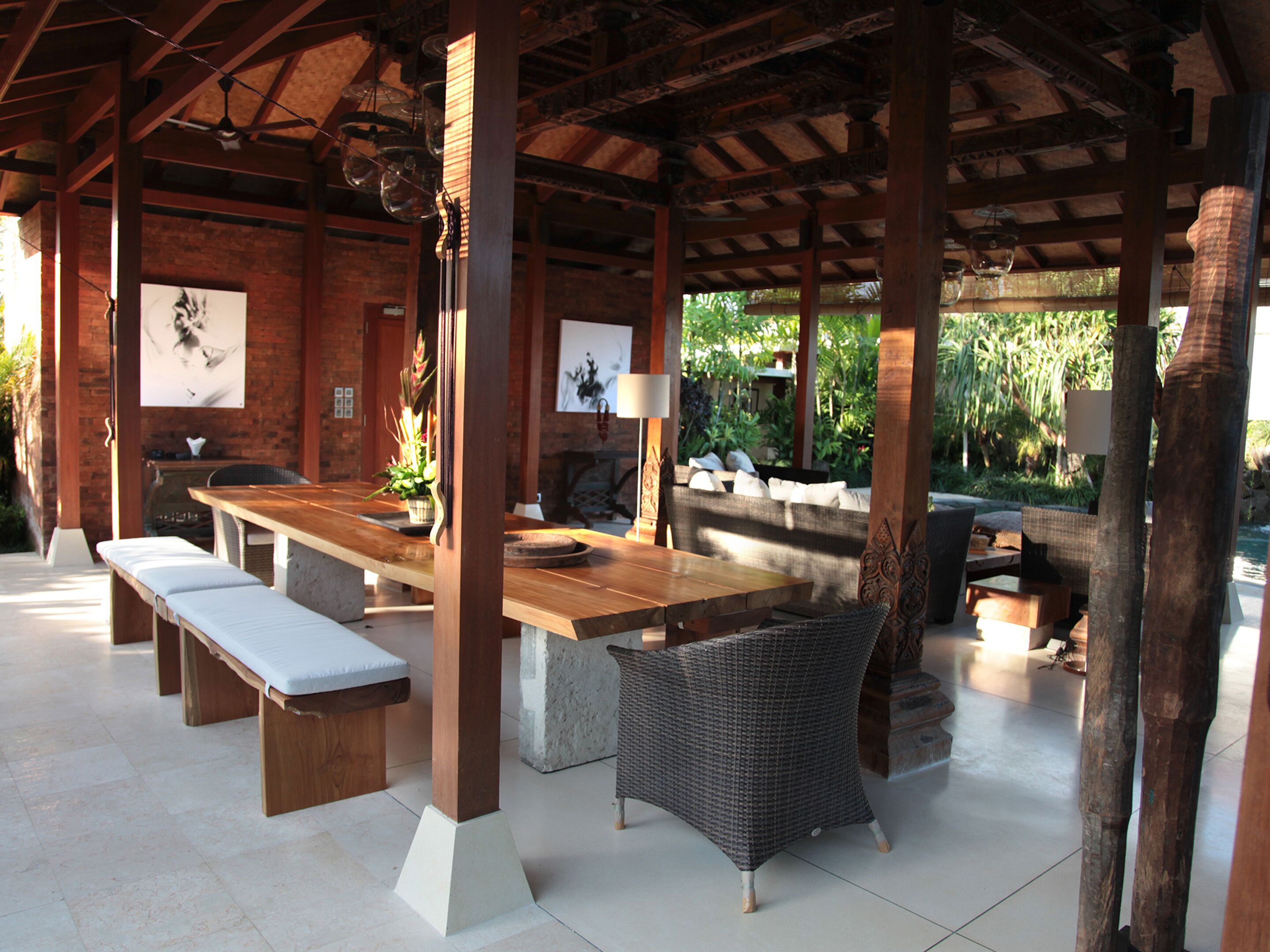 An outdoor covered patio with a wooden table, benches, wicker chairs, and a stone fireplace.