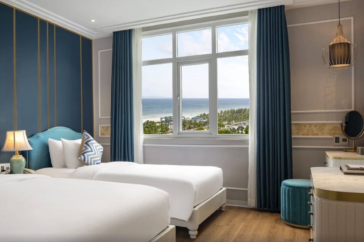 A hotel room with two beds, a large window with a view of the sea, a desk with a lamp, and a mirror.