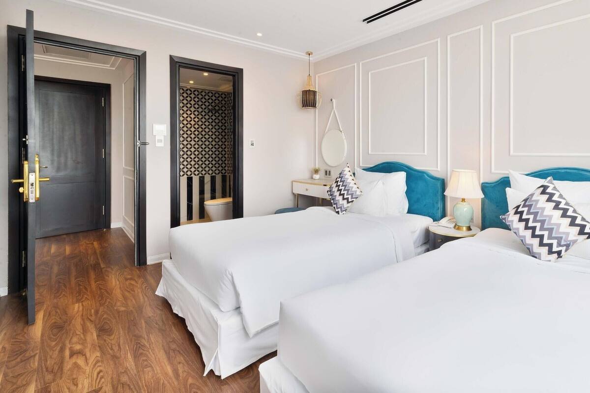 A hotel room with two beds, a wooden floor, a bathroom with a patterned door, and a nightstand with a lamp.