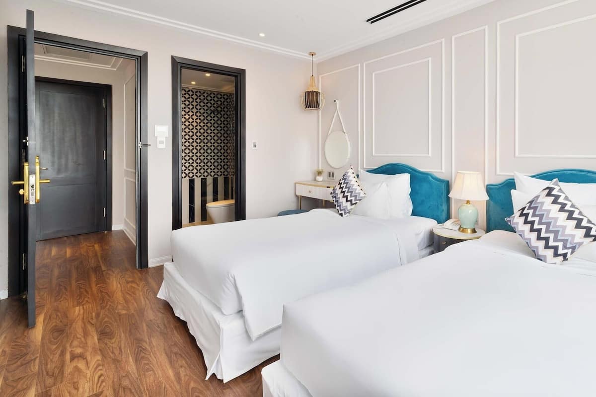 Una habitación de hotel con dos camas, suelo de madera, baño con puerta decorada y mesita de noche con lámpara.