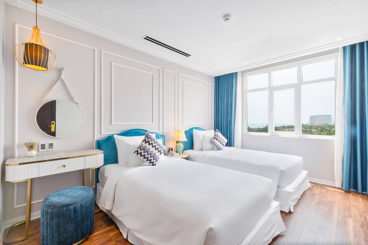 A hotel room with two beds, a vanity with a mirror, a desk, and a window with curtains.