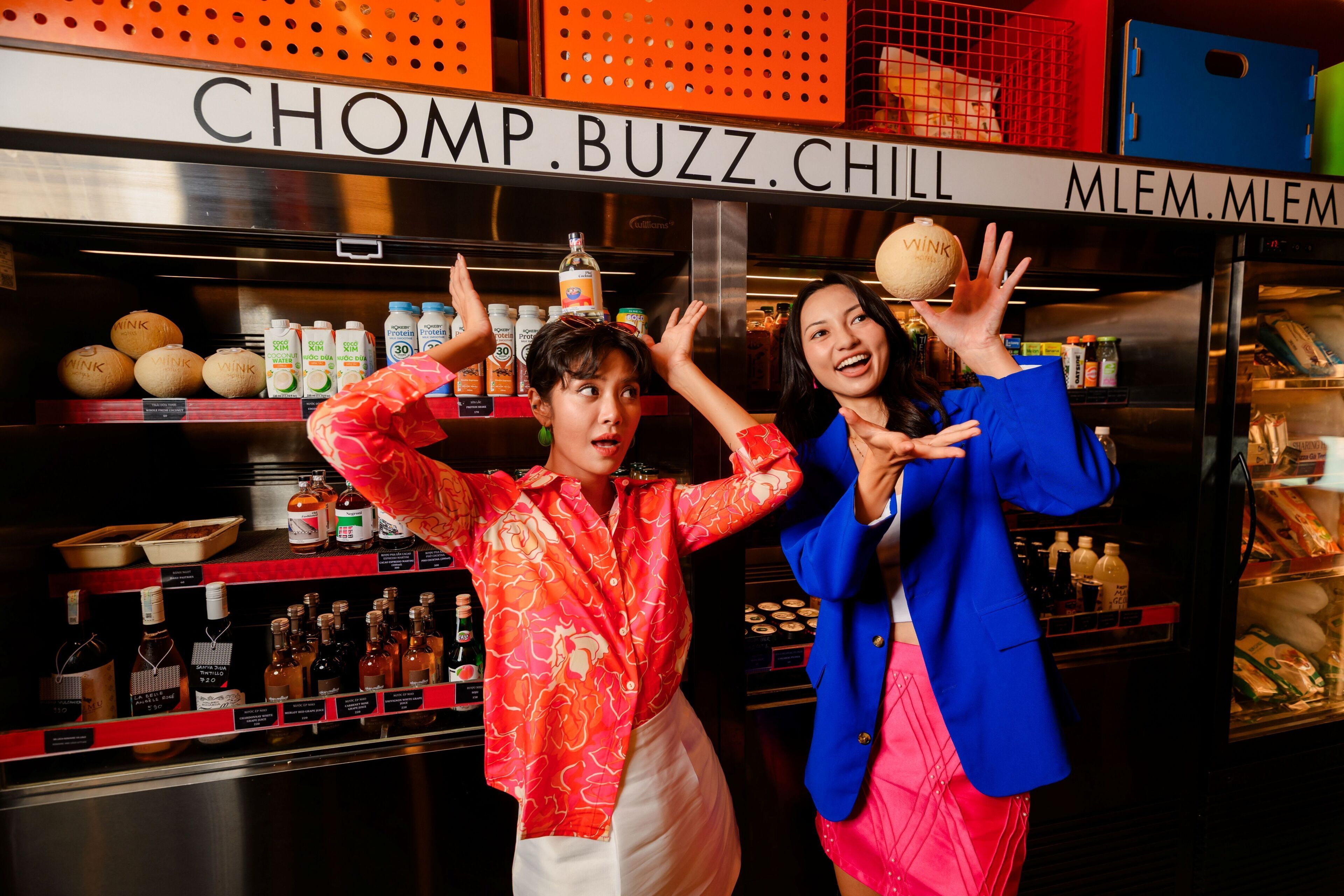 Two individuals in a bar setting with shelves stocked with bottles and a sign reading 'CHOMP.BUZZ.CHILL'.