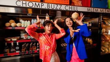 Two individuals in a bar setting with shelves stocked with bottles and a sign reading 'CHOMP.BUZZ.CHILL'.