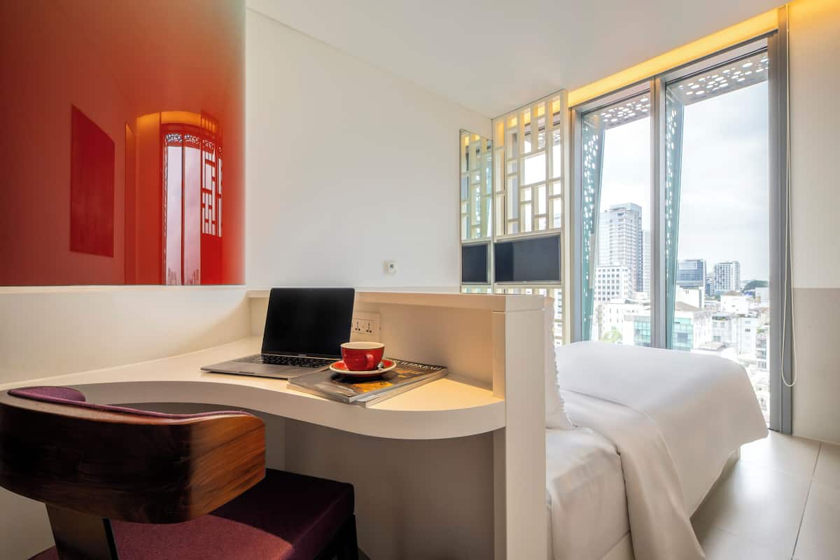 Una habitación de hotel moderna con escritorio, computadora portátil y una taza, una cama y vistas a la ciudad a través de grandes ventanales.