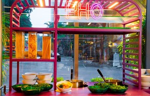 Un puesto de comida con un letrero de neón, recipientes de vidrio con fideos, tazones y una olla sobre el mostrador.