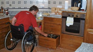 A kitchen with a person in a wheelchair, wooden cabinets, a microwave, and a stove.