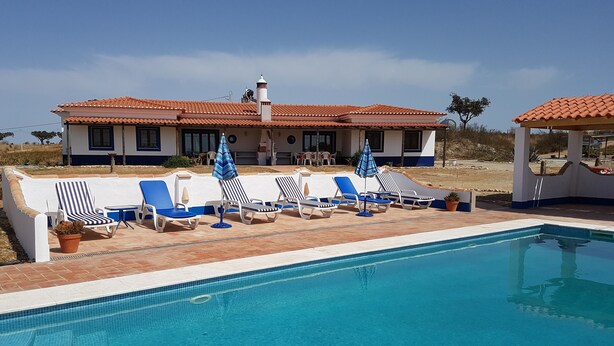 A pool area with lounge chairs and umbrellas, a tiled pool, and a building with a tiled roof in the background.