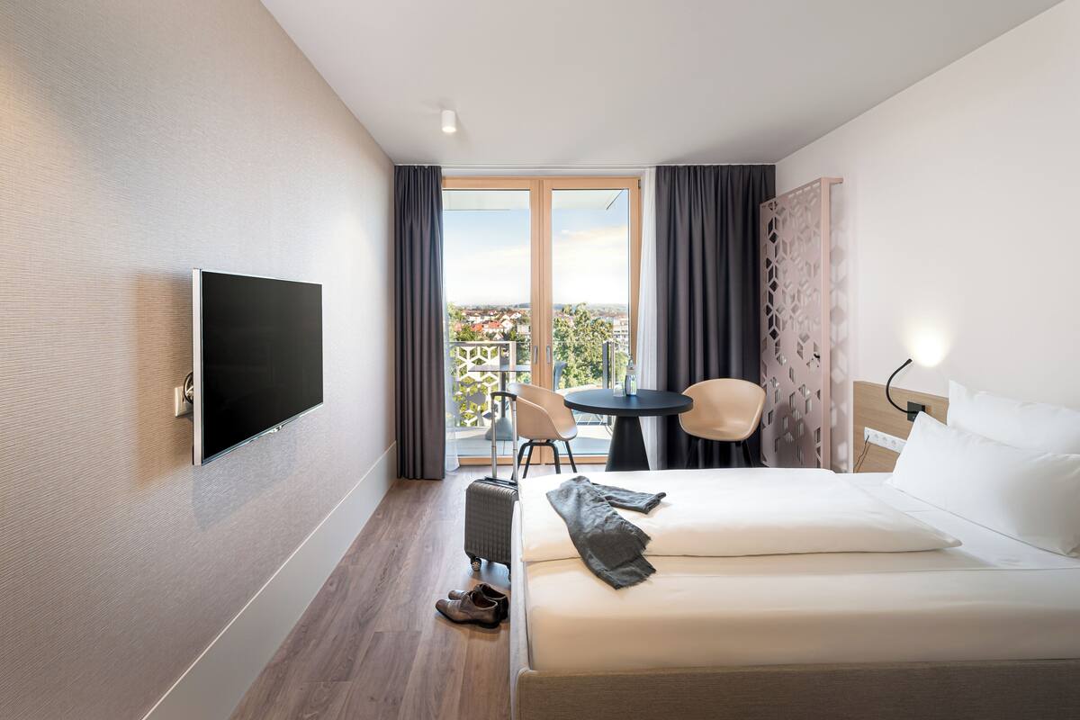 A hotel room with a bed, a TV, a desk, and a view of the city.