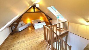 A cozy attic bedroom with a sloped ceiling, wooden beams, a bed with pillows, a chair, and a small table.