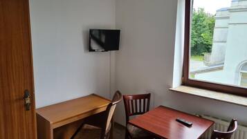A room with a wooden desk, two chairs, a television mounted on the wall, and a window with a view of greenery and a building.