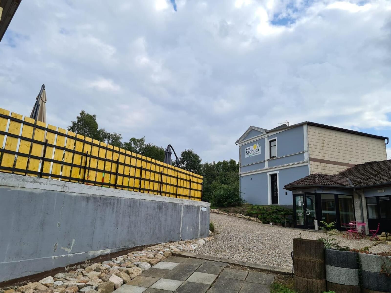 Un hôtel appelé « Hotel Seeblick », avec une façade grise, une allée gravillonnée et une clôture jaune.