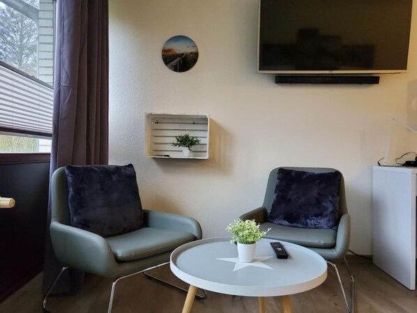 Una habitación con dos sillones, una mesita con decoración de estrella, una planta en maceta, un control remoto, un televisor montado en la pared y una ventana con persianas.