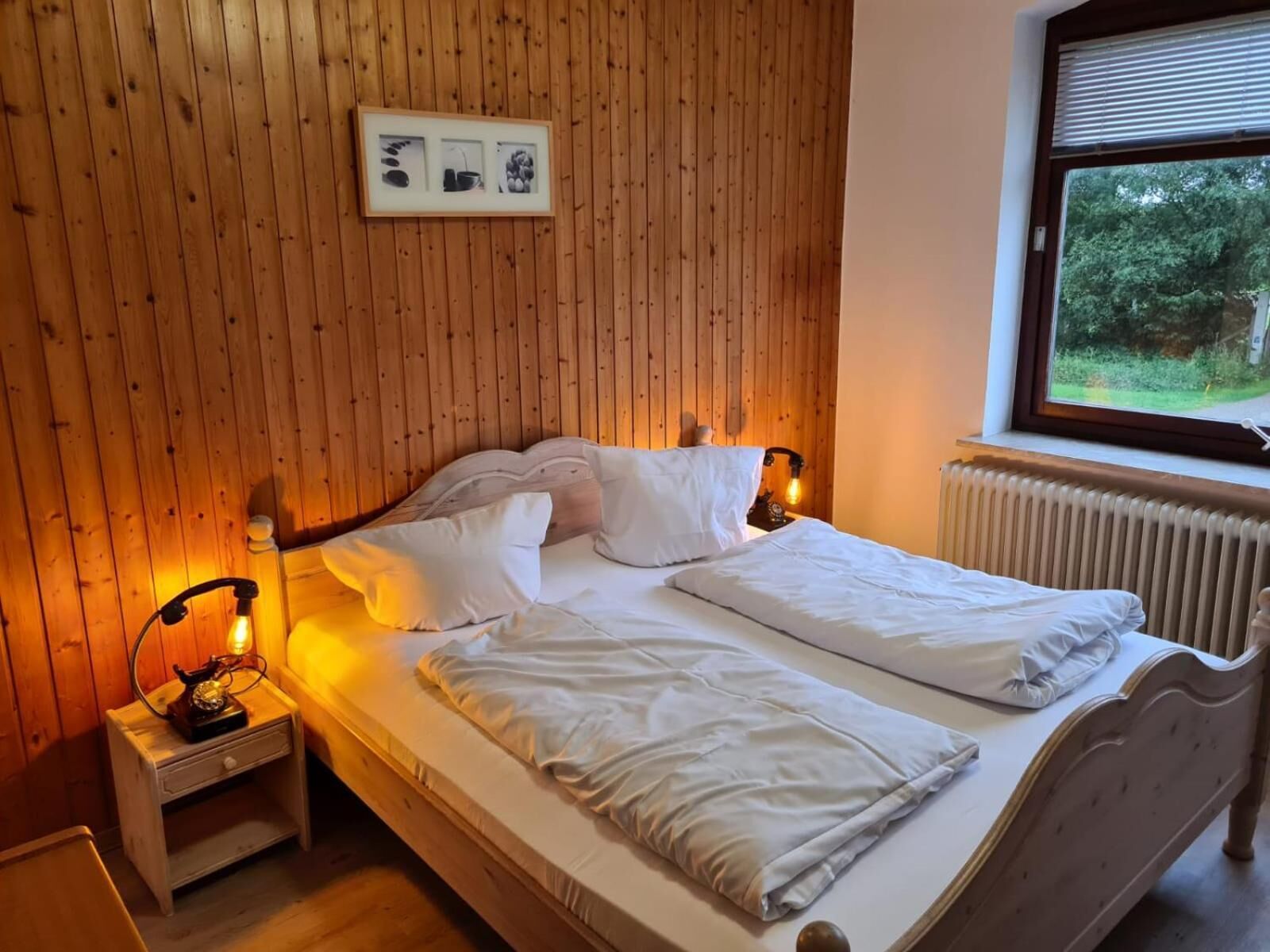 A bedroom with a wooden paneled wall, a bed with white bedding, a nightstand with a lamp, and a window with a view of greenery.