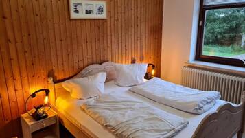 Un dormitorio con pared de madera, una cama con ropa blanca, una mesita de noche con una lámpara y una ventana con vistas a follaje.