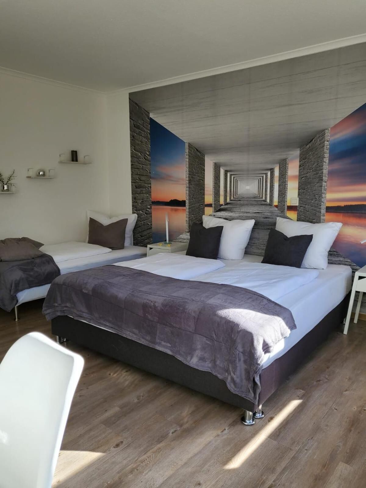 A modern hotel room with two beds, a white chair, and a view of a sunset through a window.