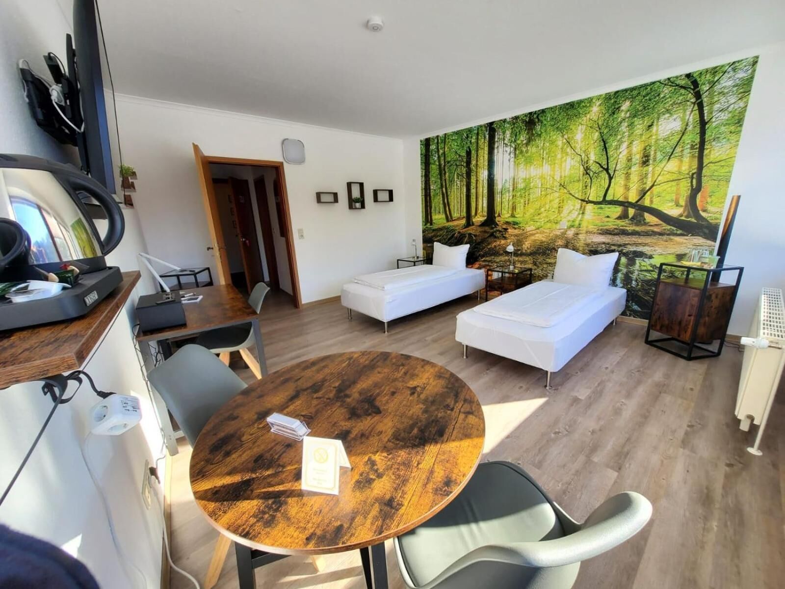 Habitació moderna amb una taula rodona de fusta, dos llits blancs, un sofà blanc i un mural gran amb un bosc a la paret.