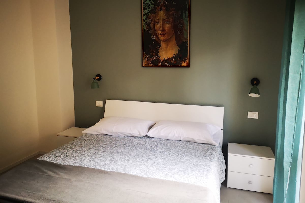 A bedroom with a bed, a nightstand, a wall-mounted lamp, and a painting.