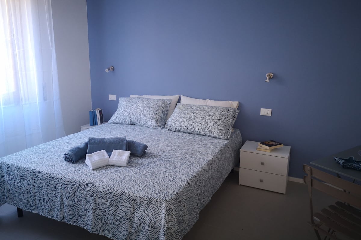 A neatly made bed with patterned bedding, a bedside table with books, and a small desk with a chair.
