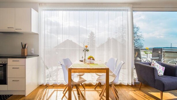 Una cocina moderna con armarios blancos, una mesa de comedor con sillas blancas y vistas al exterior a través de cortinas traslúcidas.