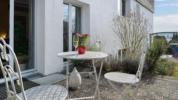 Eine Terrasse im Freien mit rundem Tisch, zwei Stühlen, einem Blumentopf und einem Fenster mit Aussicht.