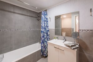 Een moderne badkamer met een bad, douchegordijn, wastafel met spiegel en een betegelde spatwand.