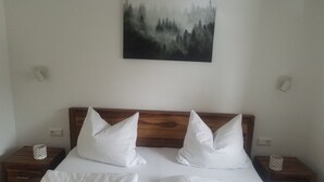 A bedroom with a wooden headboard, two white pillows, a white bedspread, and a forest-themed painting on the wall. - Quietly located vacation apartment in Pfronten (Pfronten)