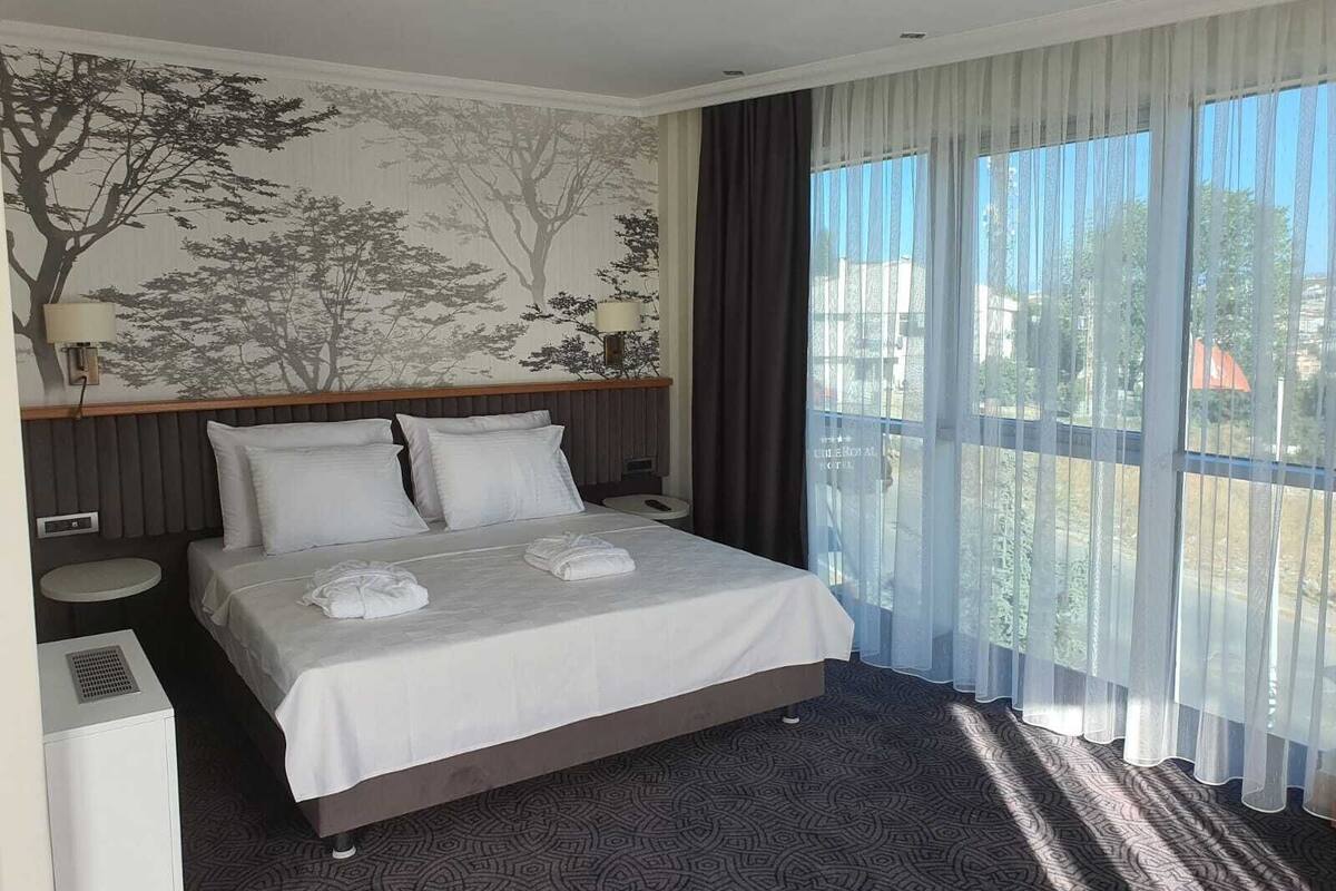 Une chambre d’hôtel avec un lit, une table de chevet ronde, une lampe fixée au mur et une fenêtre avec des rideaux légers.