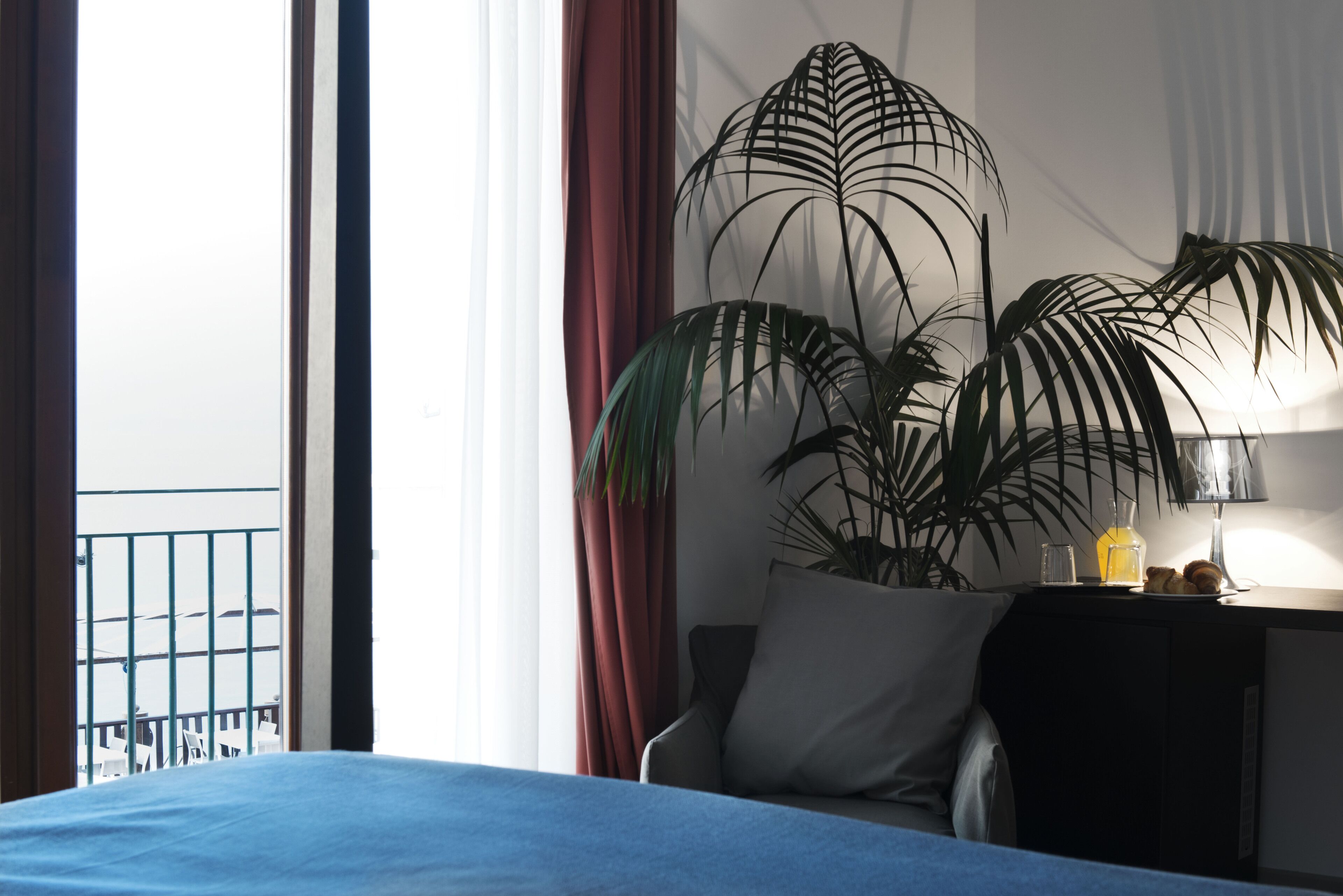 A hotel room with a bed, a nightstand, a lamp, a potted plant, and a view of the balcony.