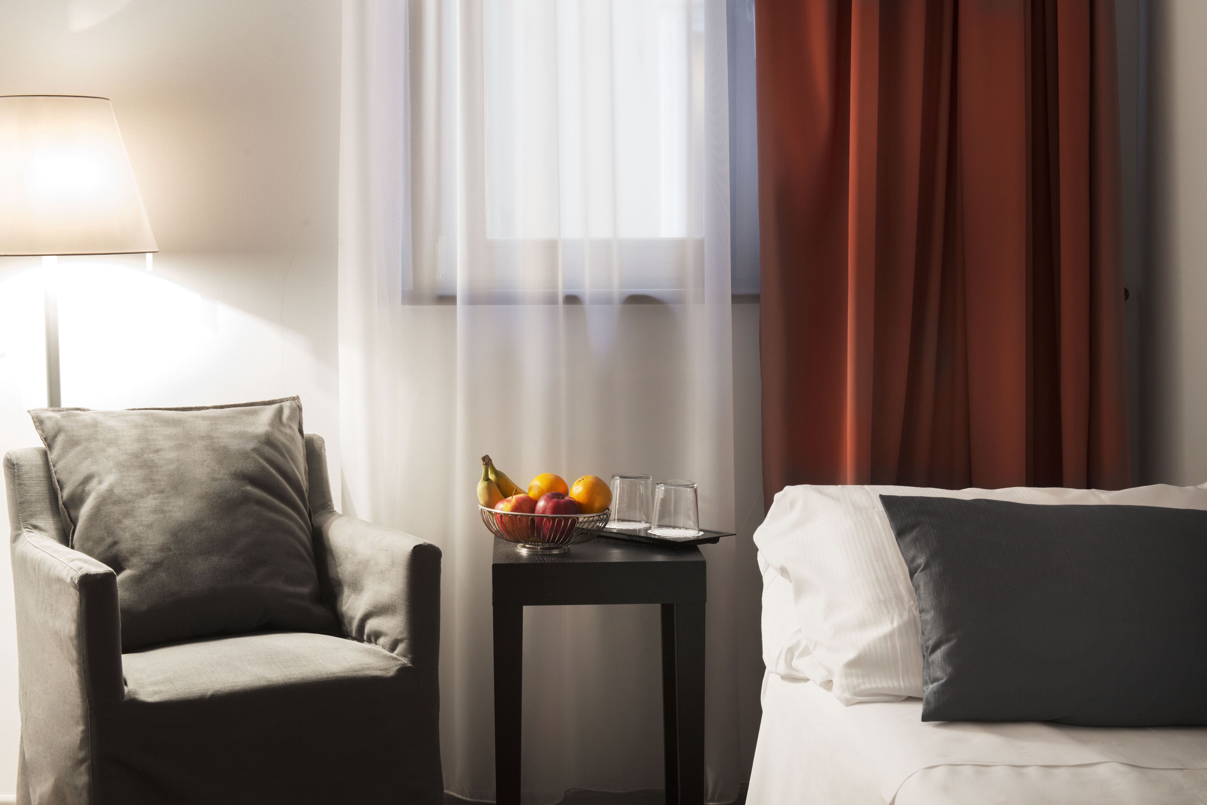 Une chambre d’hôtel avec un fauteuil gris, une petite table sur laquelle se trouve un panier de fruits et des verres, et un lit avec des oreillers blancs et gris.