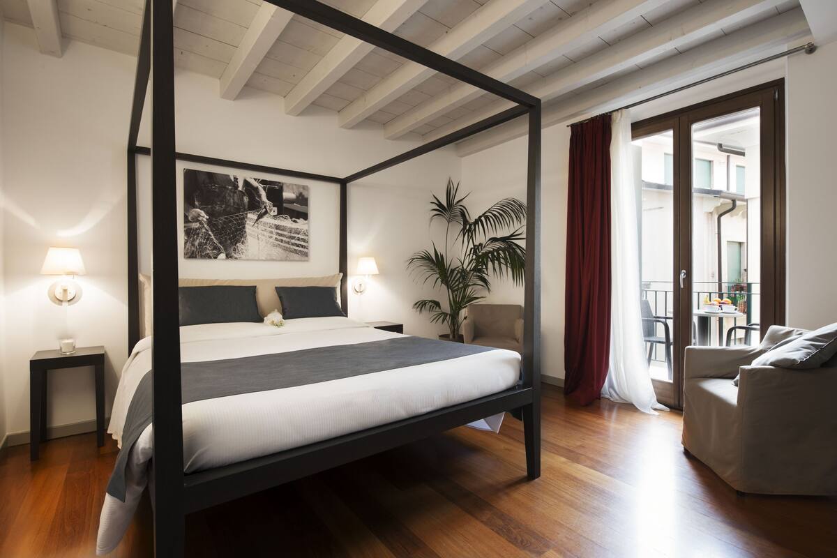 A four-poster bed with a dark frame, two bedside lamps, a large framed picture on the wall, a potted plant, and a view through a sliding glass door to an outdoor area.