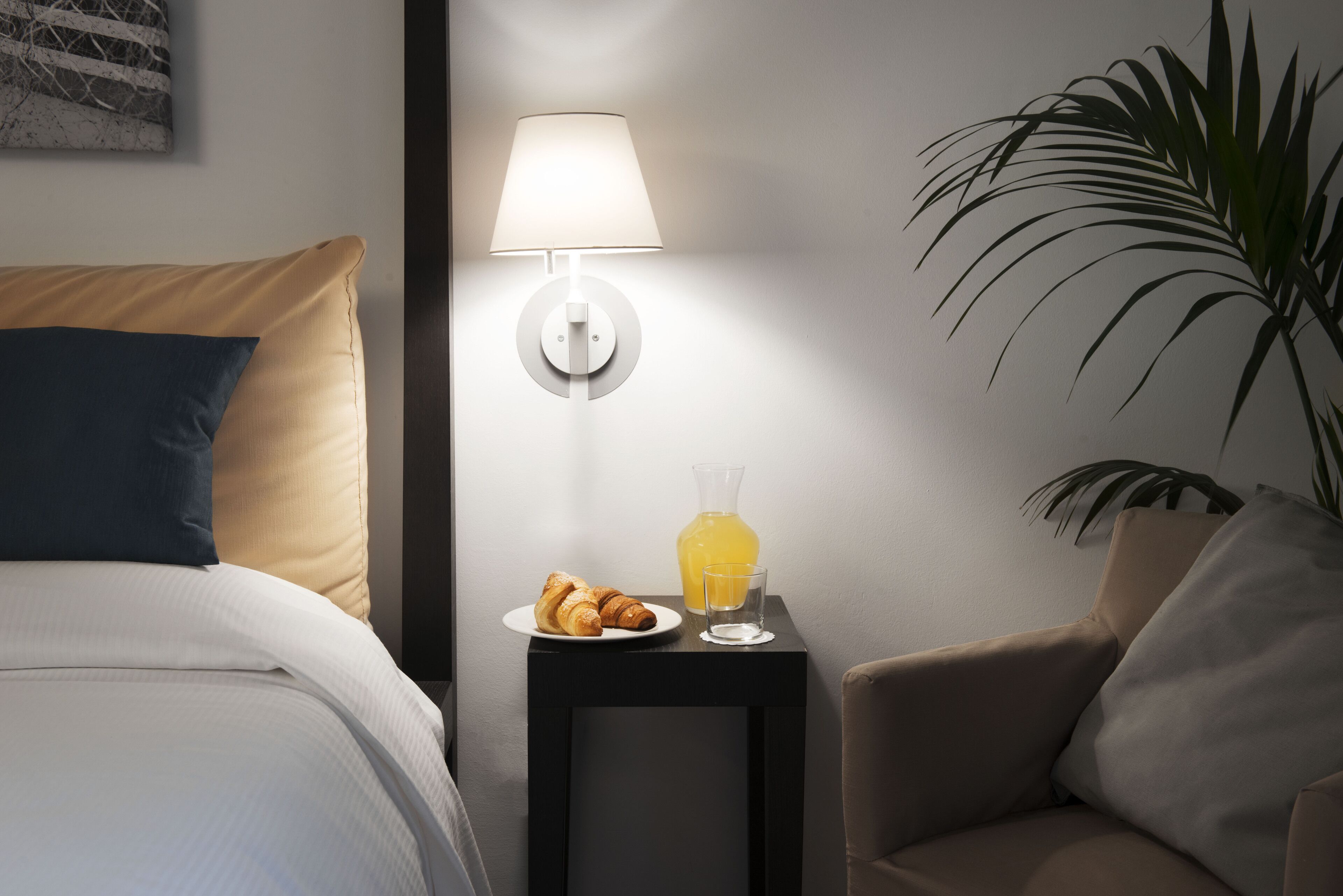 Ein ordentlich bezogenes Bett mit Kissen, ein Nachttisch mit Lampe, eine Auswahl an Croissants, ein Glas Orangensaft und ein Stuhl.