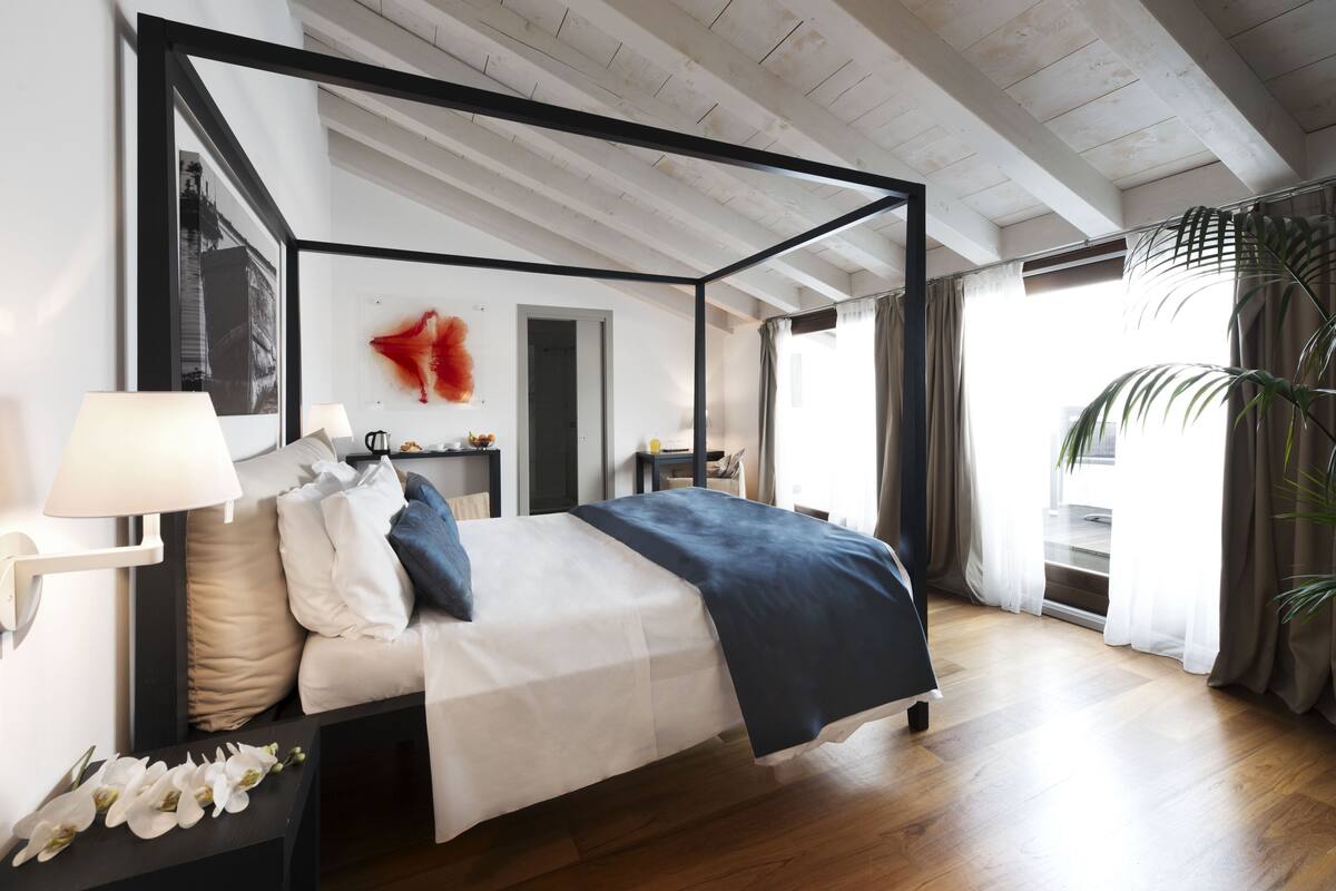 A four-poster bed with a white and blue bedspread, a bedside table with a lamp, and a wall-mounted picture frame.