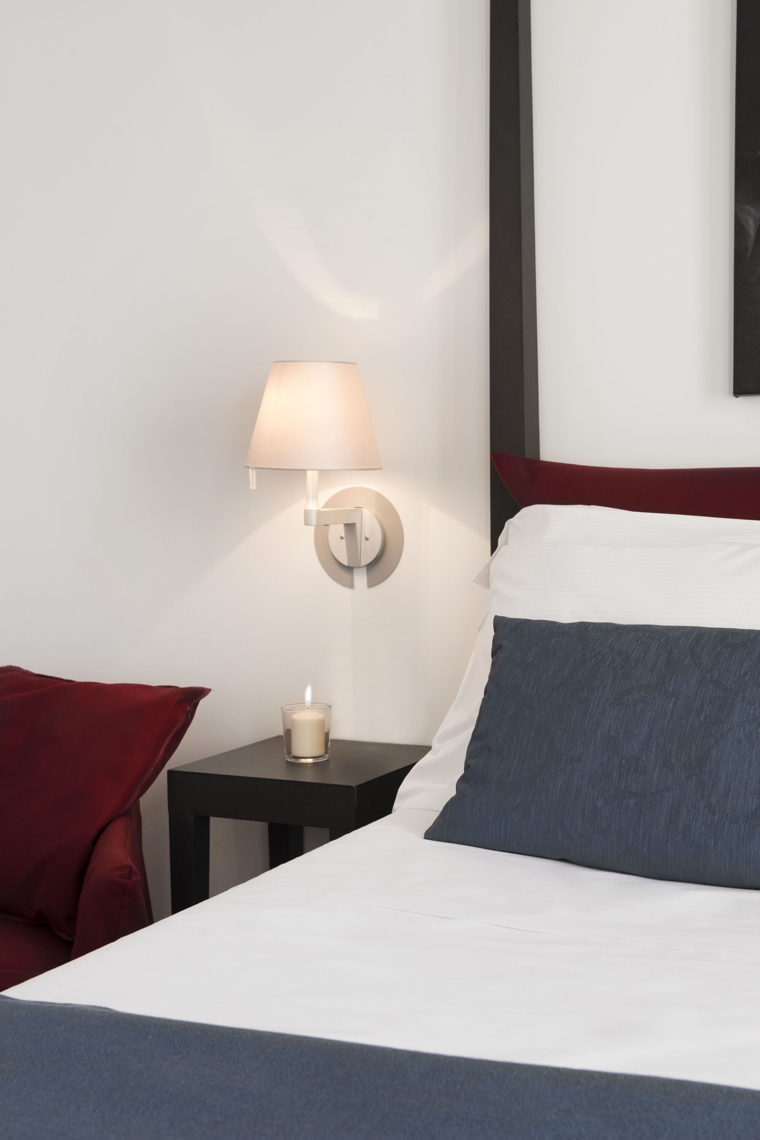 A neatly made bed with white and gray bedding, a red pillow, a small bedside table with a white lamp, and a lit candle.