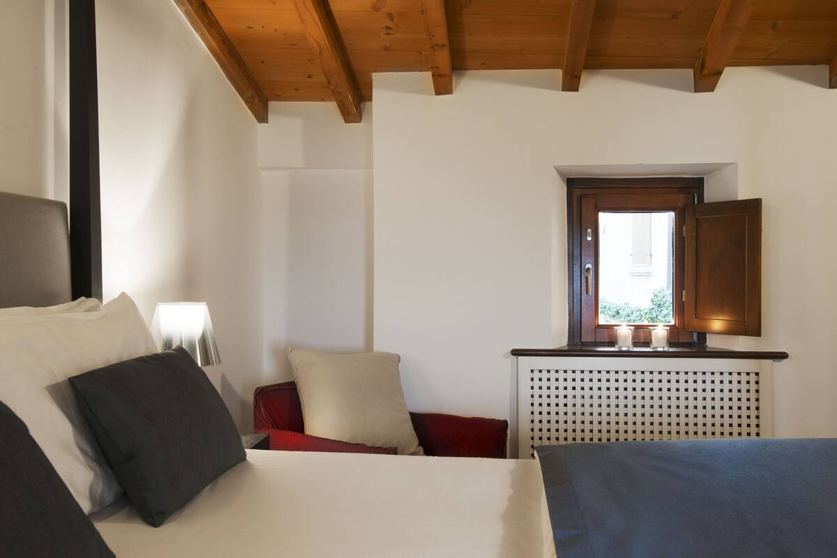 A bedroom with a bed, pillows, a nightstand, a lamp, a window, and a wooden ceiling.