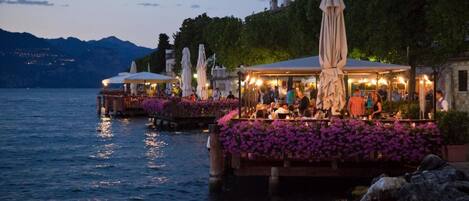 Outdoor dining areas with tables and umbrellas, overlooking a body of water with mountains in the background.