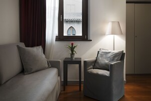Un soggiorno moderno con divano, poltrona, un tavolino con una pianta, una lampada e una finestra che offre vista su un edificio.
