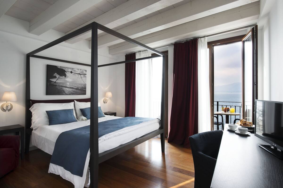 A four-poster bed with a view of the sea, a dining table with breakfast items, and a TV on a desk.