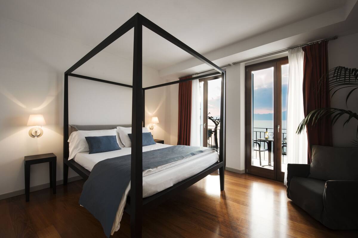 A modern bedroom with a four-poster bed, a small table with a lamp, and a view of the outdoors through a sliding glass door.