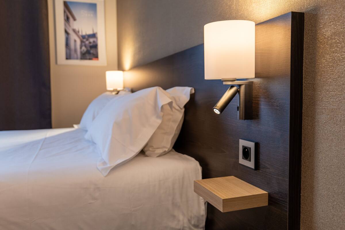 Un lit bien fait, avec du linge de lit blanc, une table de chevet avec une lampe et un luminaire fixé au mur.