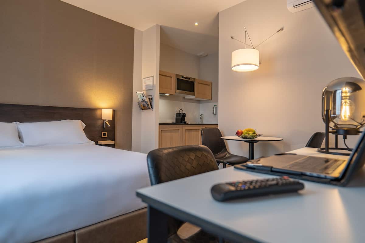 Une chambre d’hôtel avec un lit, un bureau avec un ordinateur portable, une chaise, une télécommande, une lampe, une petite table avec des fruits, et, au fond, une kitchenette.